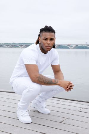 Athletic figure in all-white outfit crouches on wooden dock by waterfront.