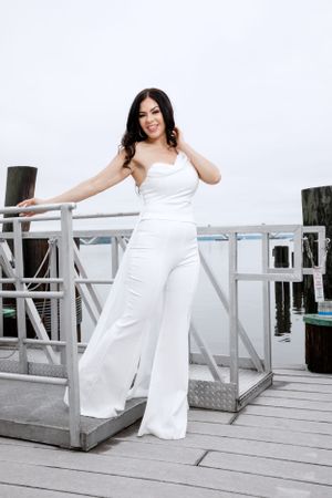 Woman in white jumpsuit poses cheerfully on dock railing near water.