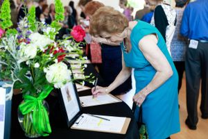 Person in turquoise dress signing documents next to floral arrangements at an event.