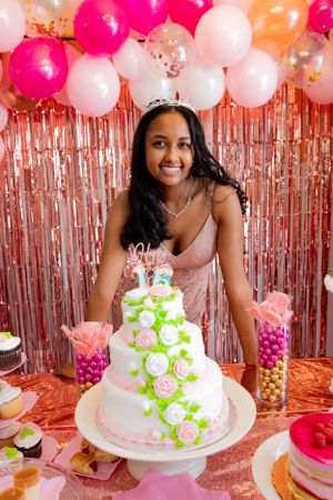 A beautifully decorated white and pink tiered cake with balloons and streamers in the background.