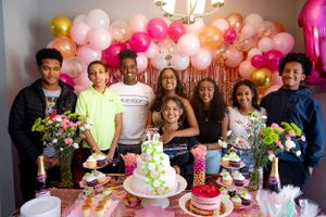 Group gathered around a tiered cake at a celebration with pink and gold balloon decorations.