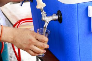 Hand dispensing a beverage from a blue drink cooler into a clear plastic cup.