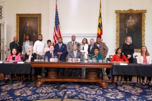 Group photo of Governor Wes Moore, legislators and officials seated at long table in government chamber with state flags.