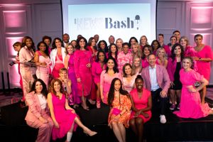 Large group of News Anchors wearing pink and formal attire poses together at a Bash event with pink lighting.