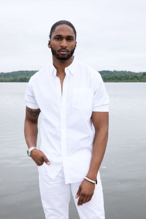 Man in crisp white shirt and pants stands confidently on dock by the water.