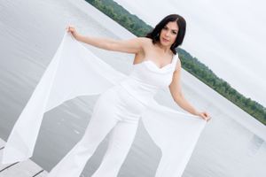 Woman in flowing white jumpsuit poses gracefully on dock by the water.
