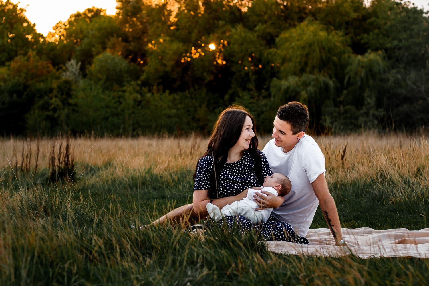New parents sit in a field in Havant, Hampshire smiling at each other while holding their newborn baby boy