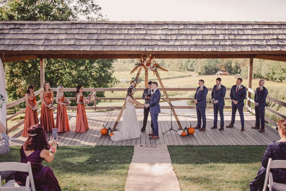 Outdoor wedding ceremony under covered wooden pavilion with bridal party in rust-colored dresses.