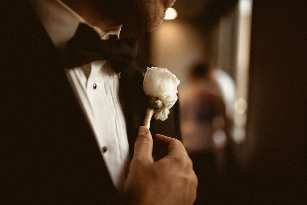 Close-up of groom adjusting white boutonniere in dark lighting.