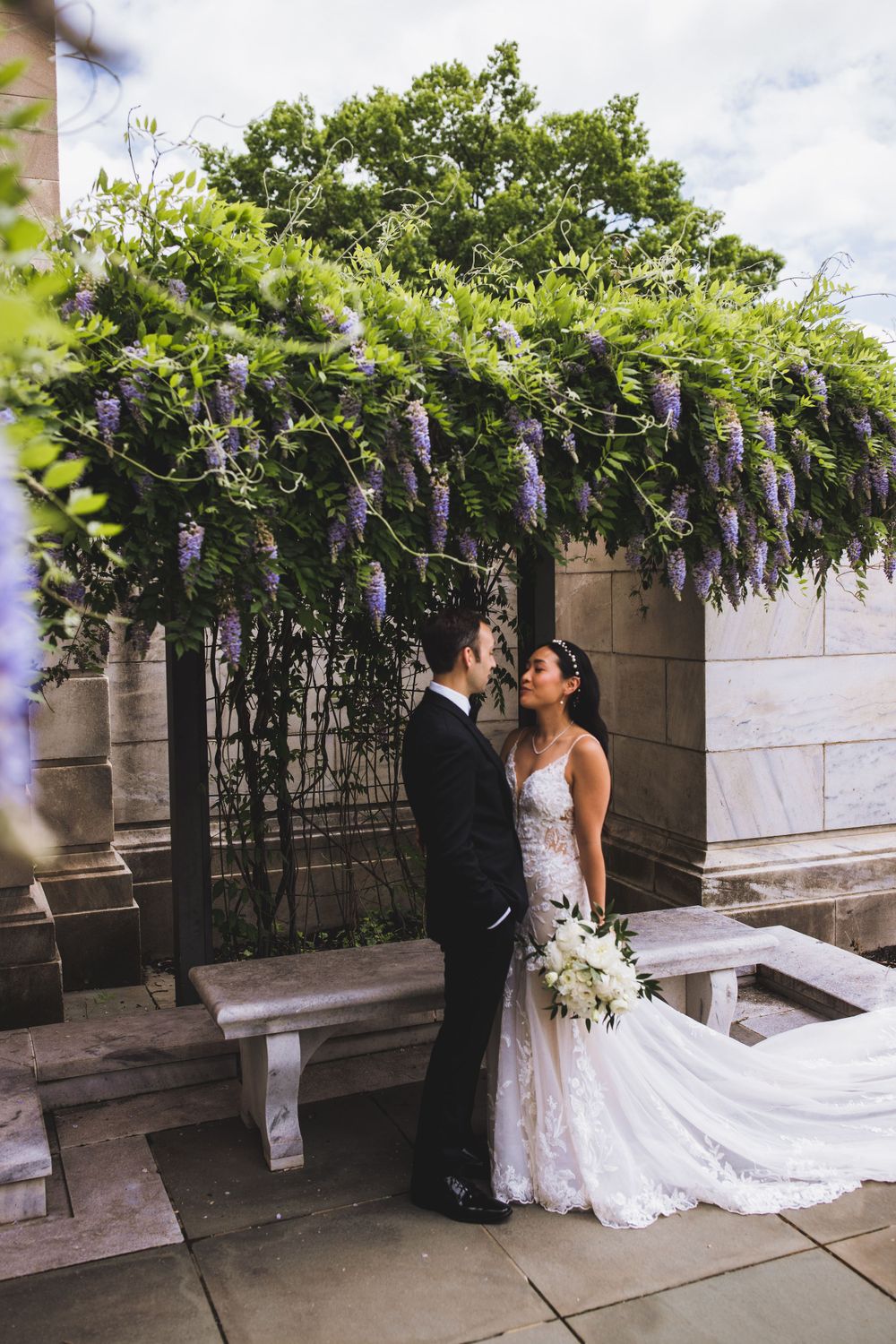 Couple in wedding attire poses beneath cascading purple wisteria flowers near stone architecture.