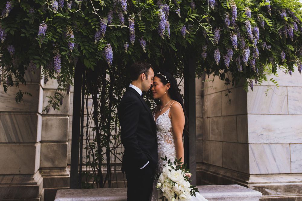 Newlywed couple embraces under purple wisteria flowers against stone wall with white bouquet.