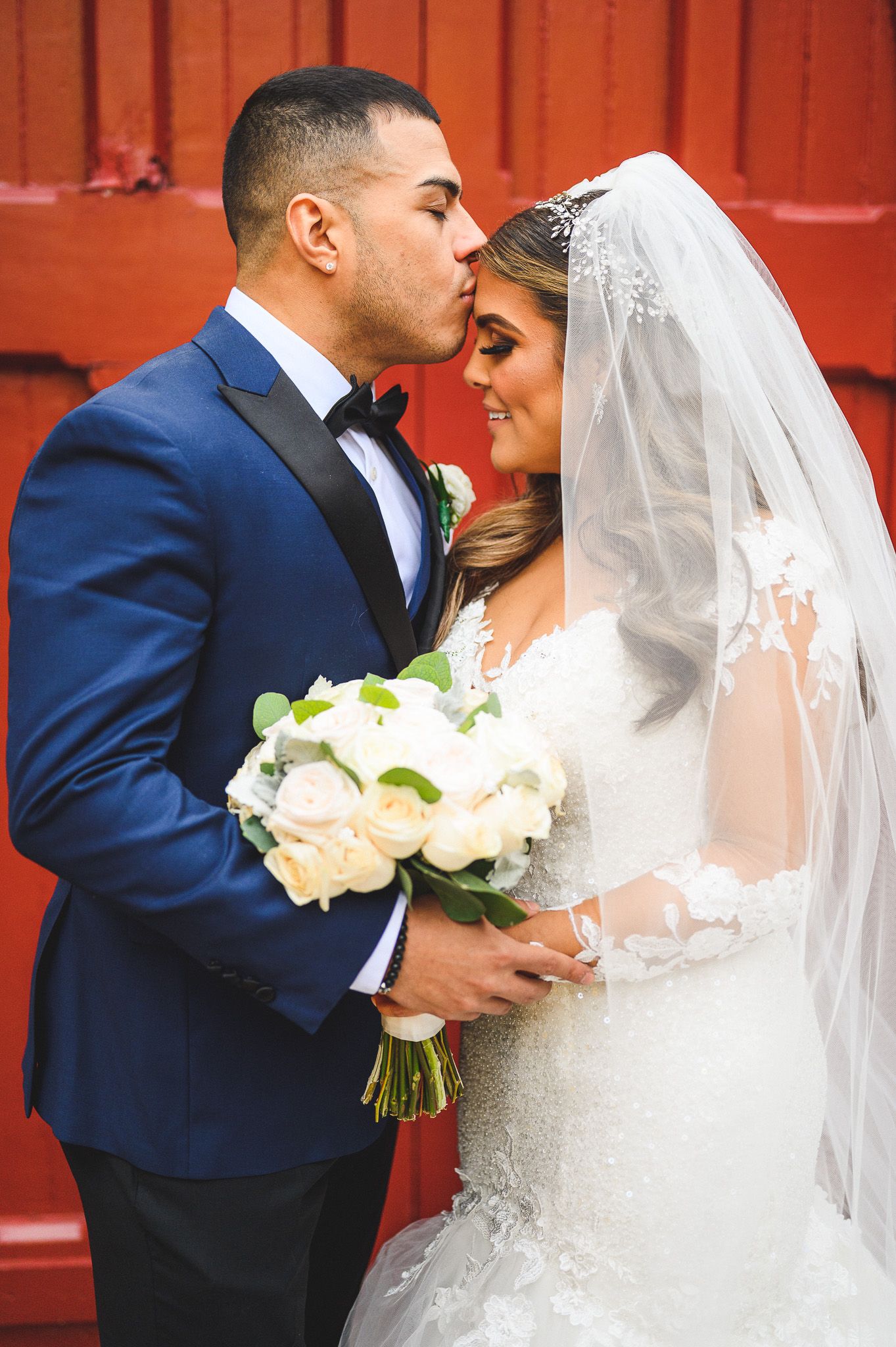 groom kissing his bride forehead in front of red doors at stone house