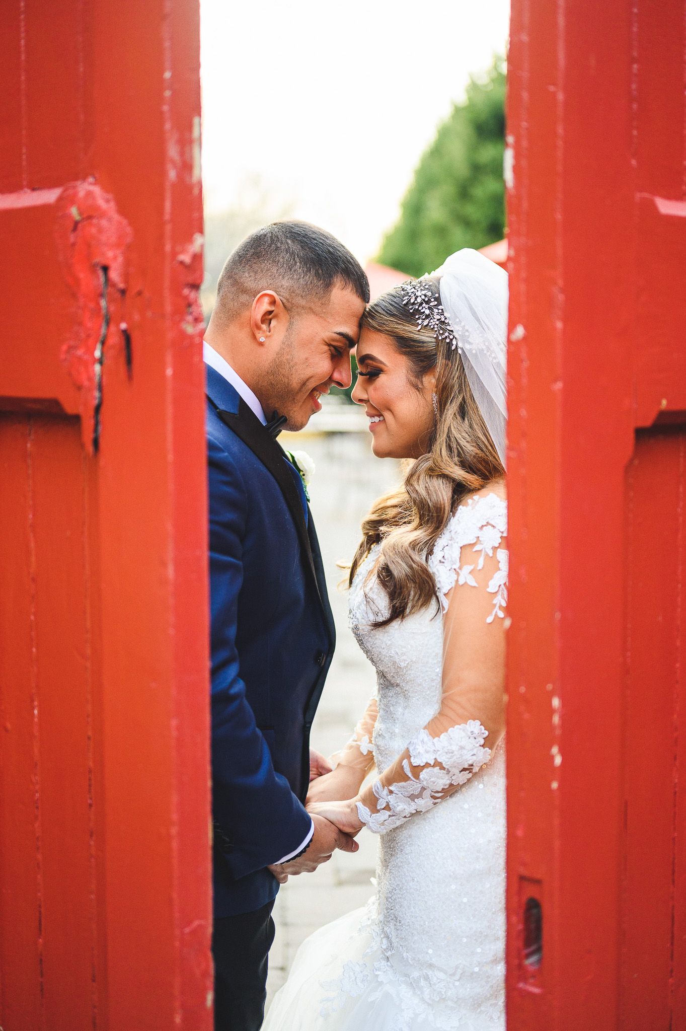 bride and groom smiling with foreheads together between red doors at stone house