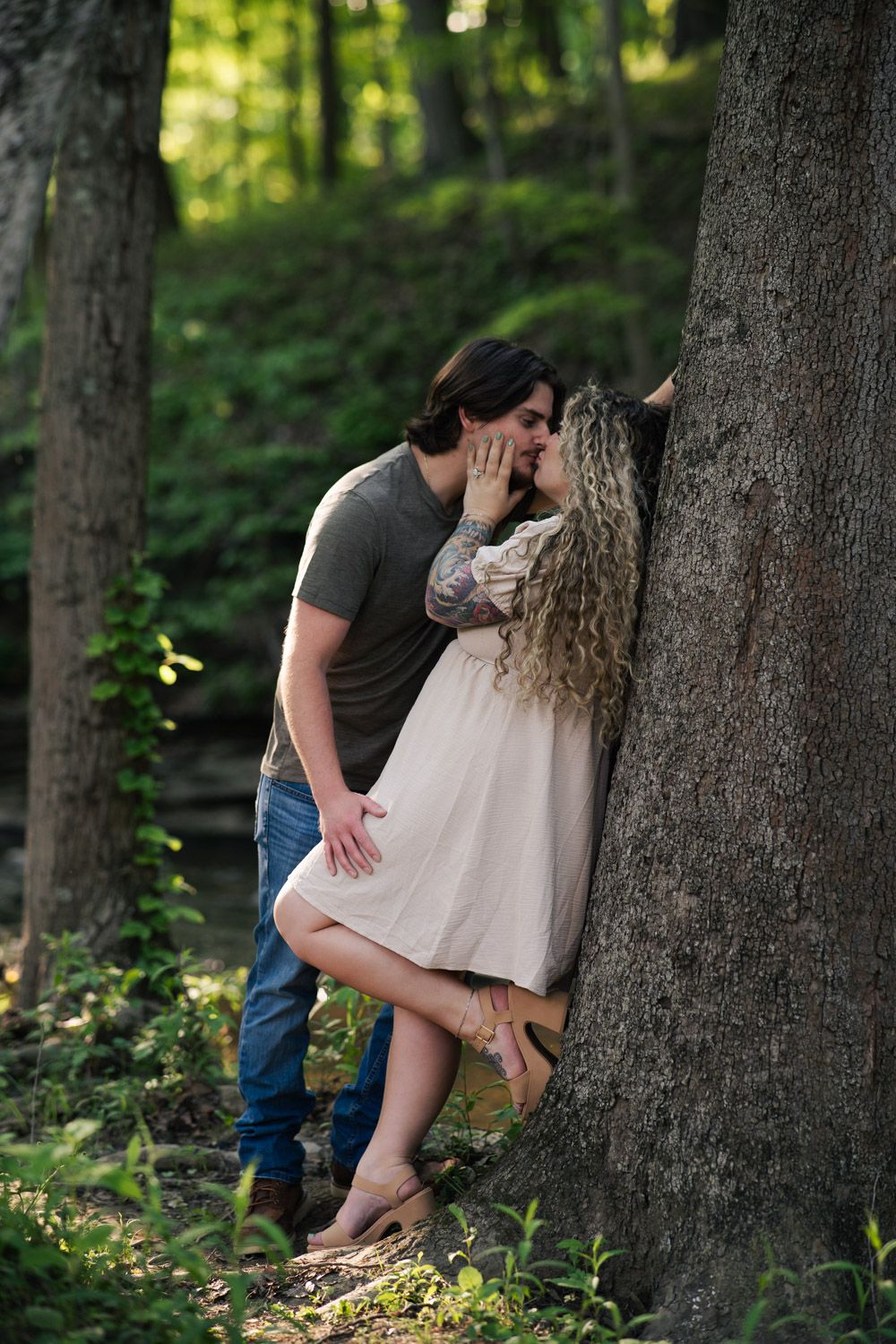 A romantic couple share an intimate moment against a tree trunk in a sunlit forest setting.