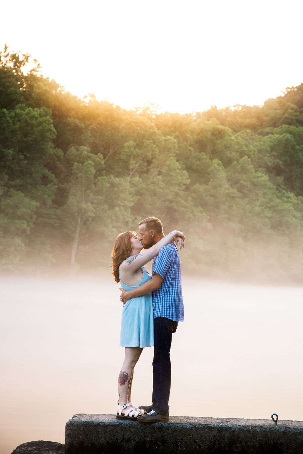 A romantic couple shares tender moments in the misty golden light beside a lake as the sun sets in the background.