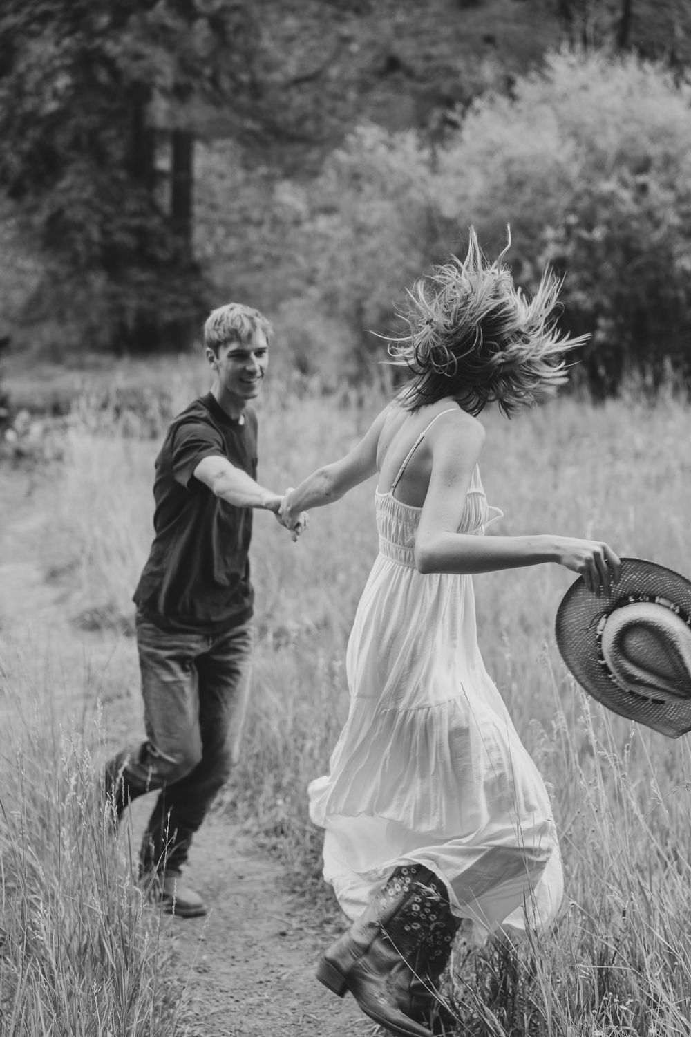 Black and white photo of a couple dancing joyfully in a meadow, with her dress flowing and hair in motion as they spin.