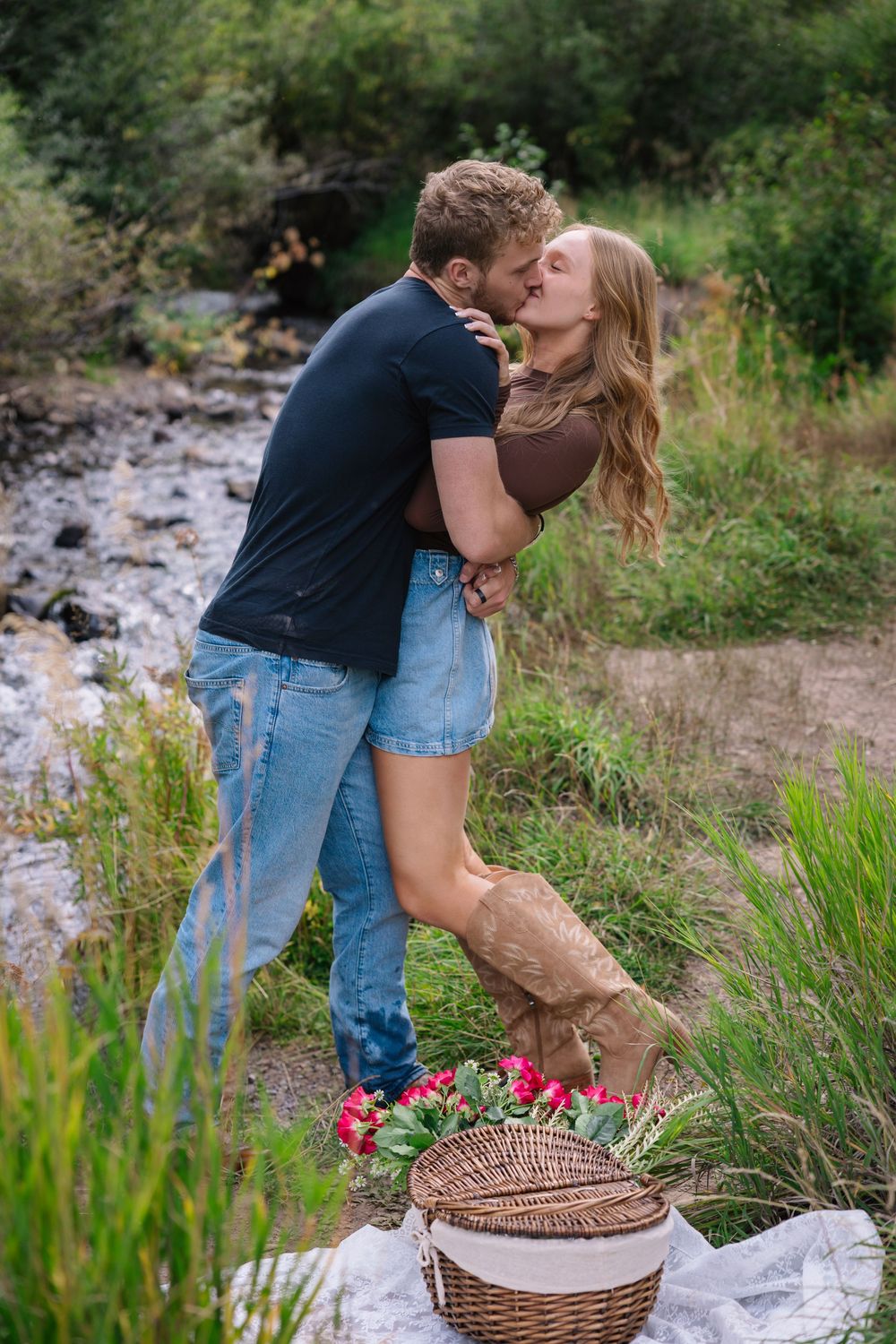 A couple shares a romantic moment by a picnic basket on a grassy riverside while surrounded by wildflowers.
