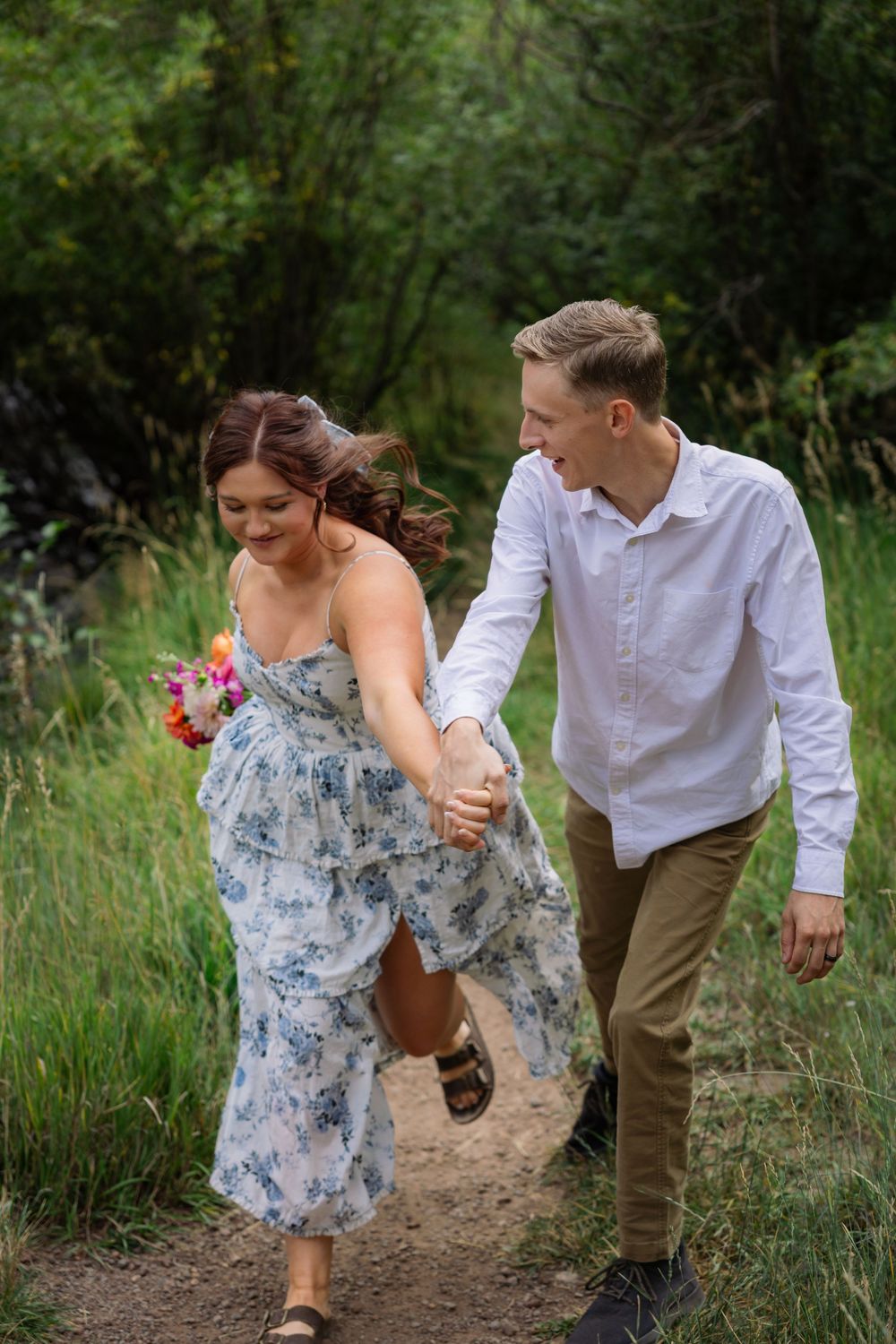 A couple walking together on a nature trail, with her wearing a floral dress and holding flowers while he wears a white shirt.
