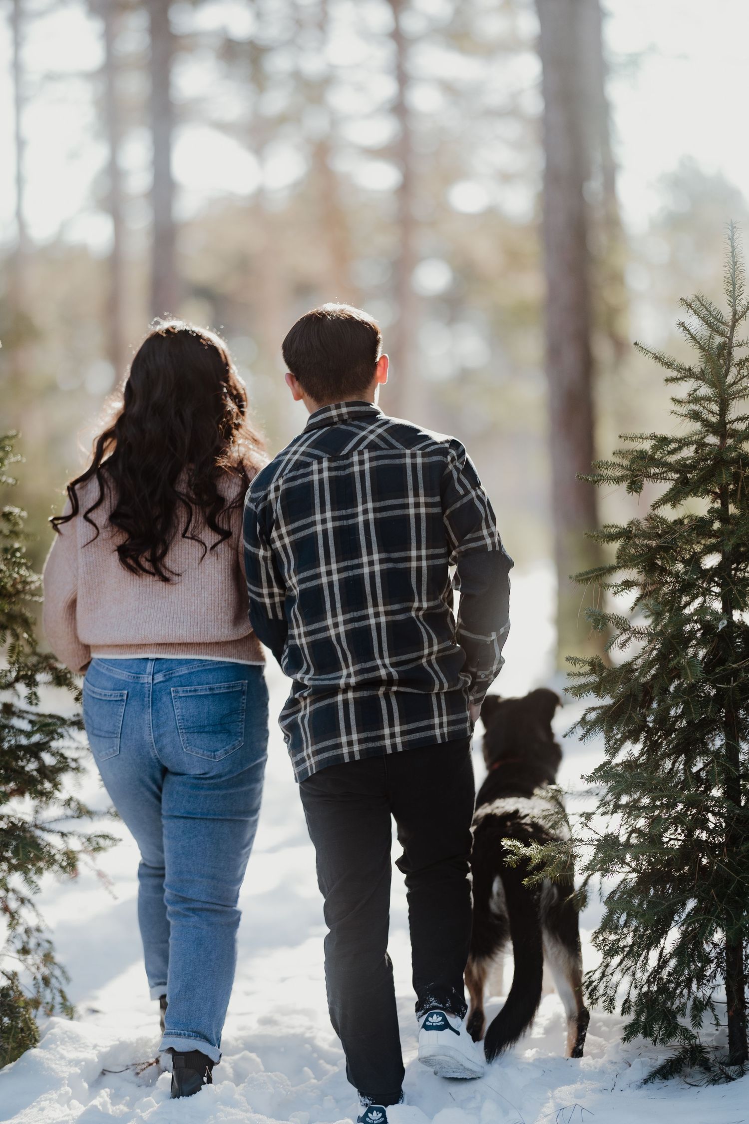 Couple walks with their dog through a snowy forest path between evergreen trees.