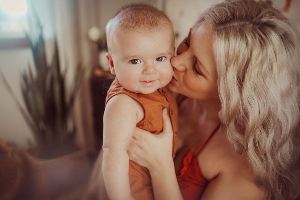 Baby in orange romper being embraced against warm interior setting.