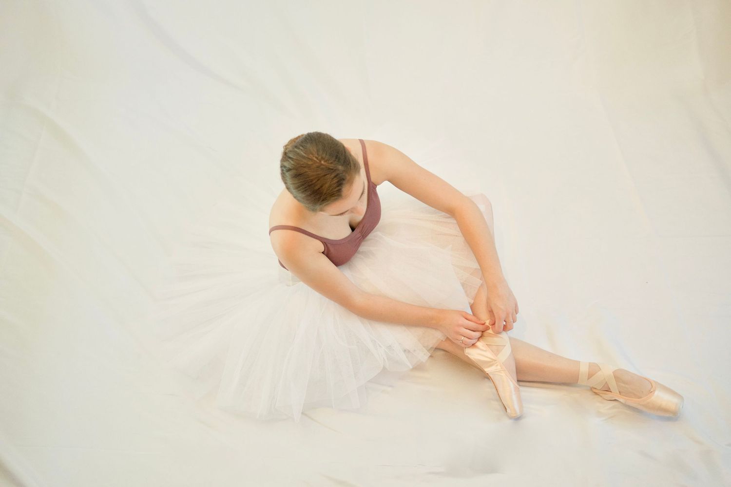 Ballerina in white tutu adjusts pointe shoes while seated on floor in elegant pose.