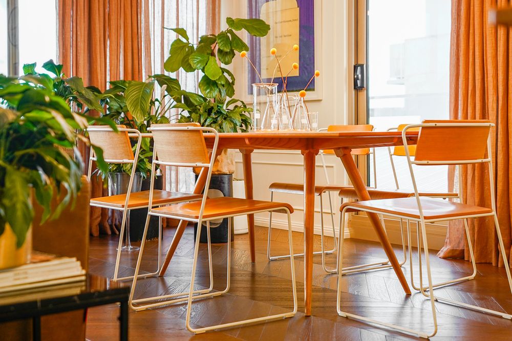 Bright dining area with orange chairs, wooden table, and lush green plants by large windows.