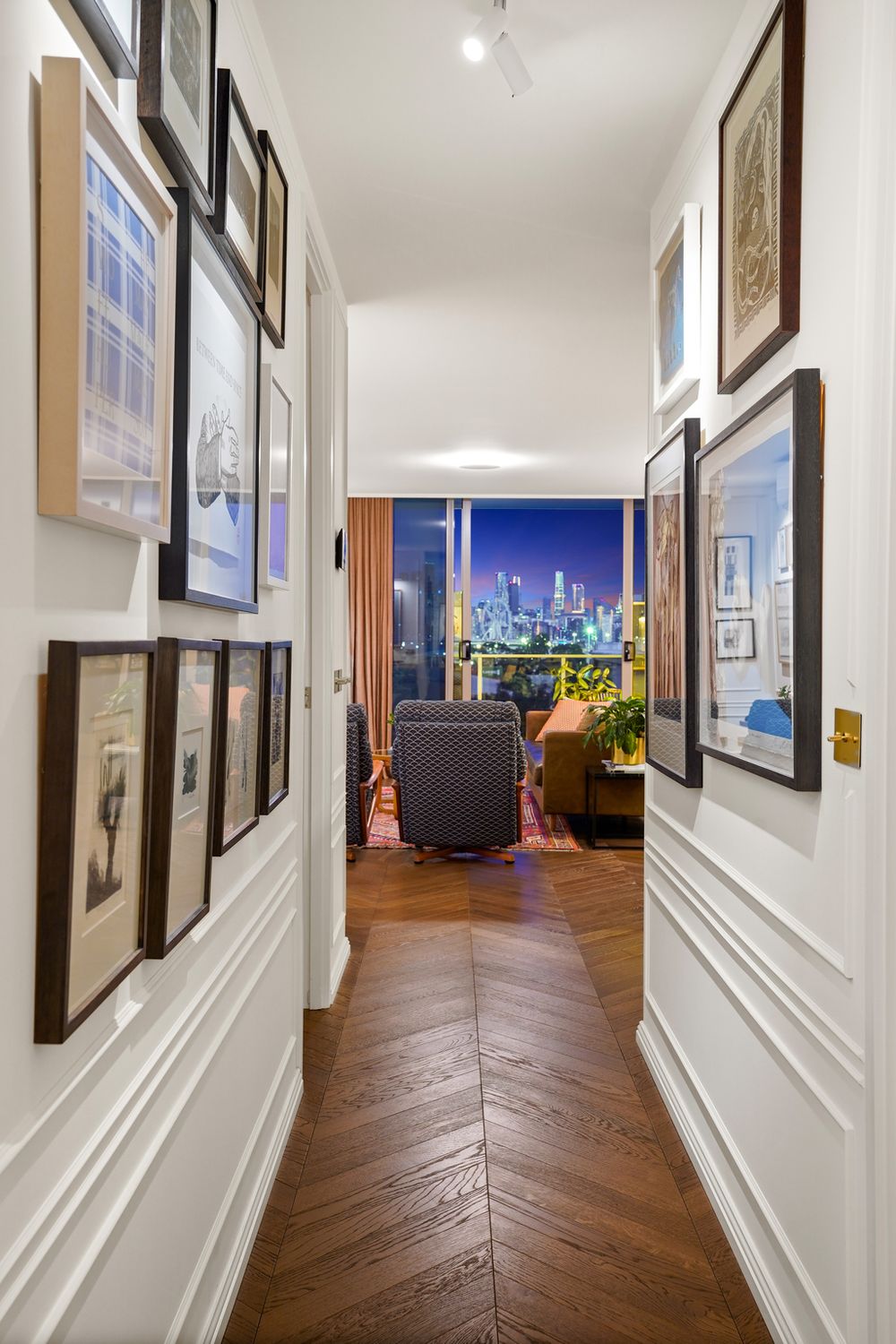 Long hallway with herringbone wood floors and gallery wall leading to city view through orange curtains.