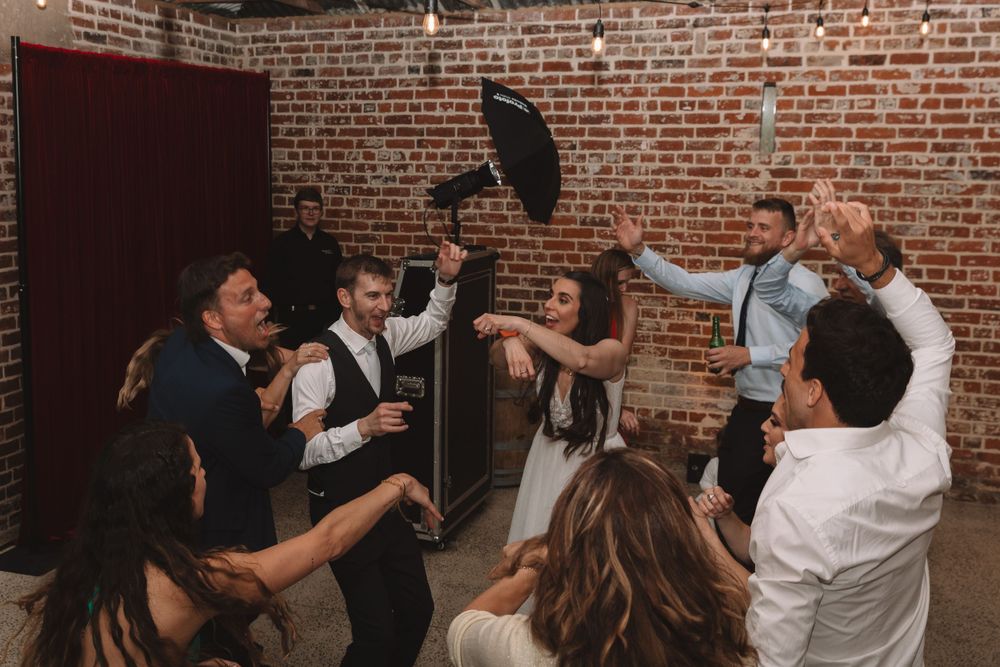 Wedding party celebrates energetically on dance floor in brick-walled venue.