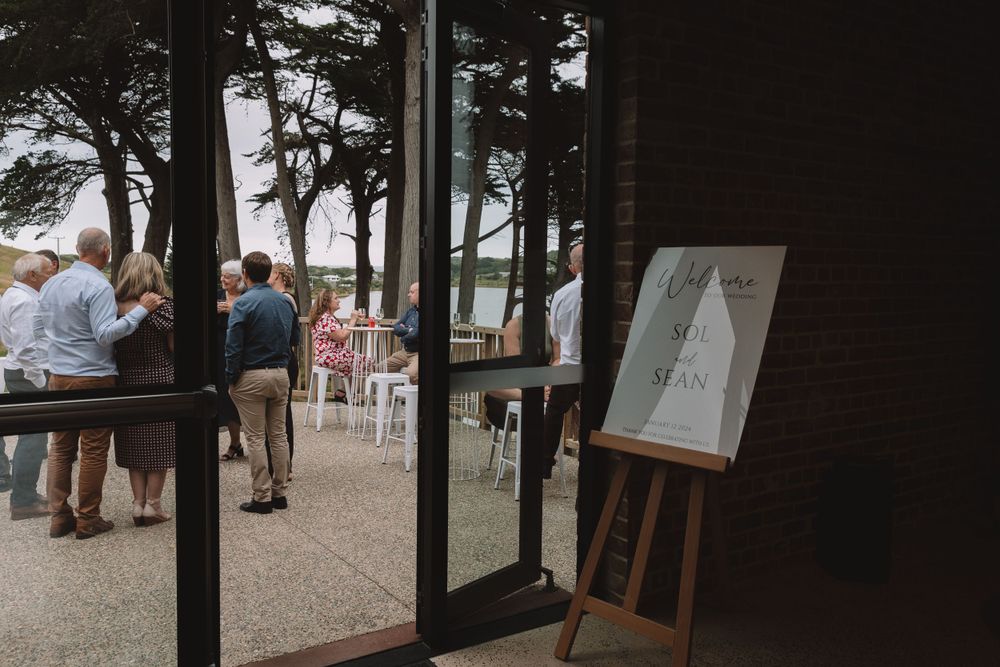 Wedding venue entrance framed by glass doors with welcome sign and pine trees in background.
