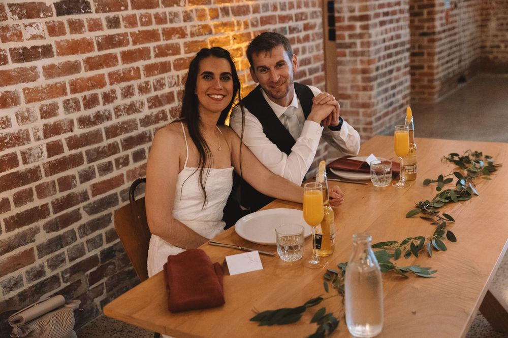 Couple enjoying refreshments at rustic reception with exposed brick walls and floral decor.