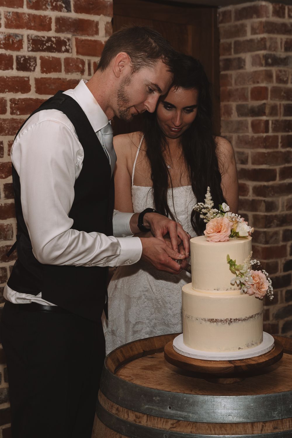 A couple cutting their elegant white wedding cake adorned with flowers on a wooden barrel.
