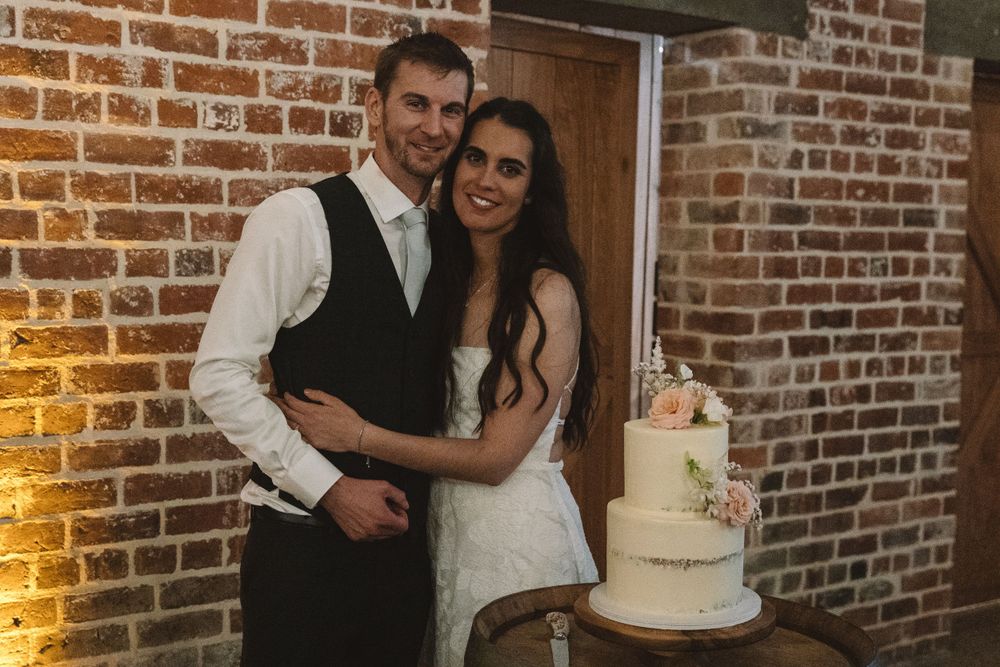 A couple posing with their white wedding cake on display against a rustic brick wall.