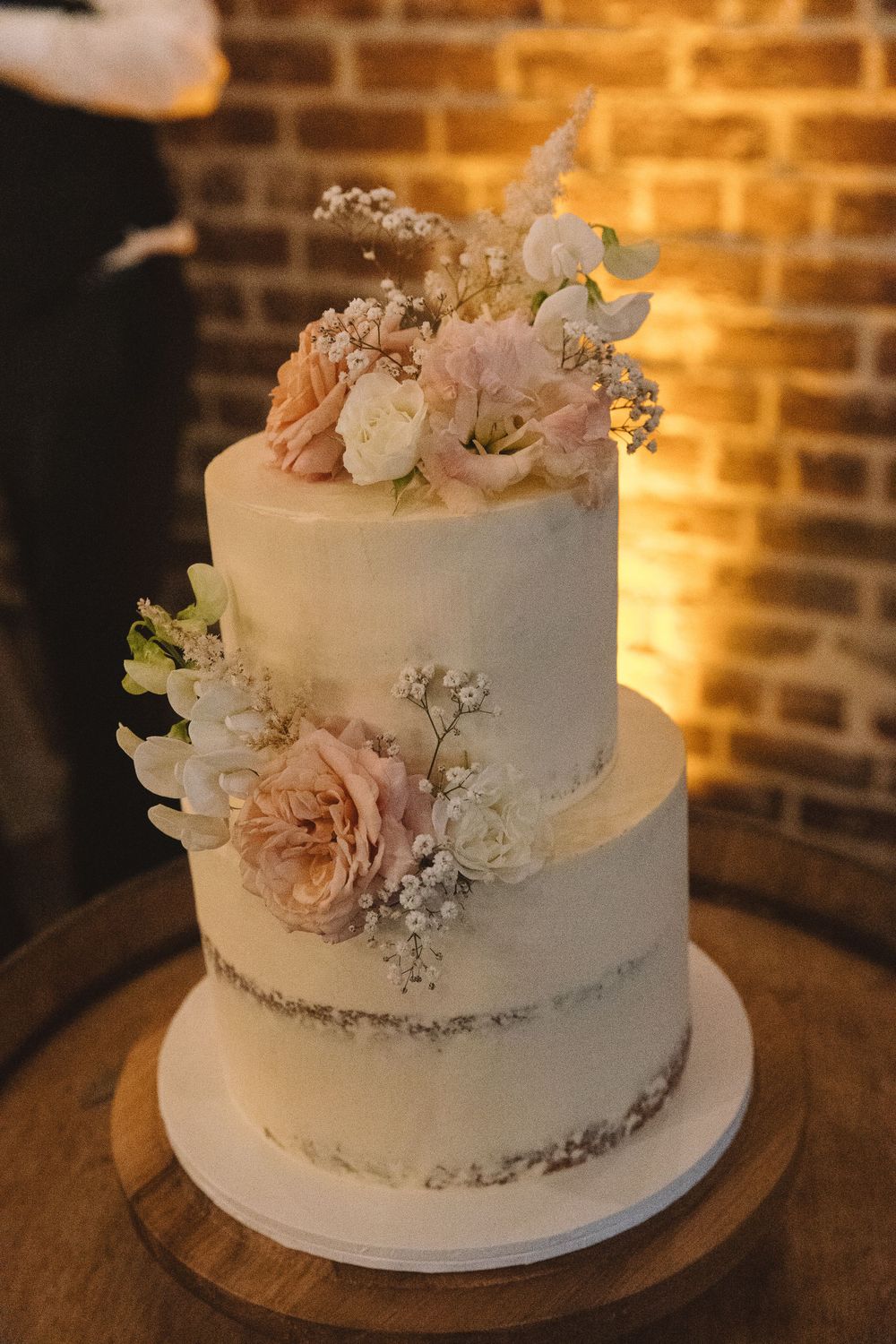 Two-tiered white wedding cake decorated with blush roses and baby's breath on wooden barrel stand.