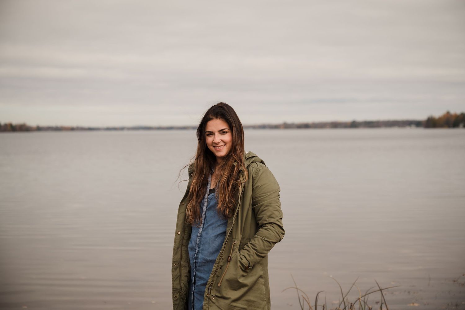 Woman in green jacket stands by calm lake on overcast day.