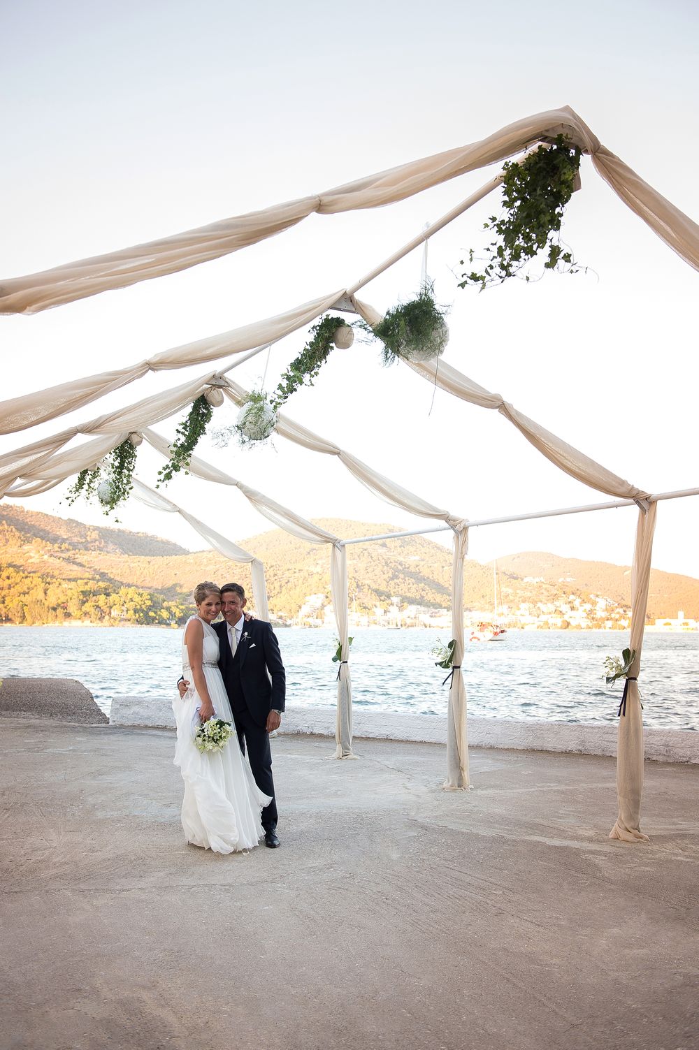 couple posing in front of the poros sea under a chapel decorated with green leaves