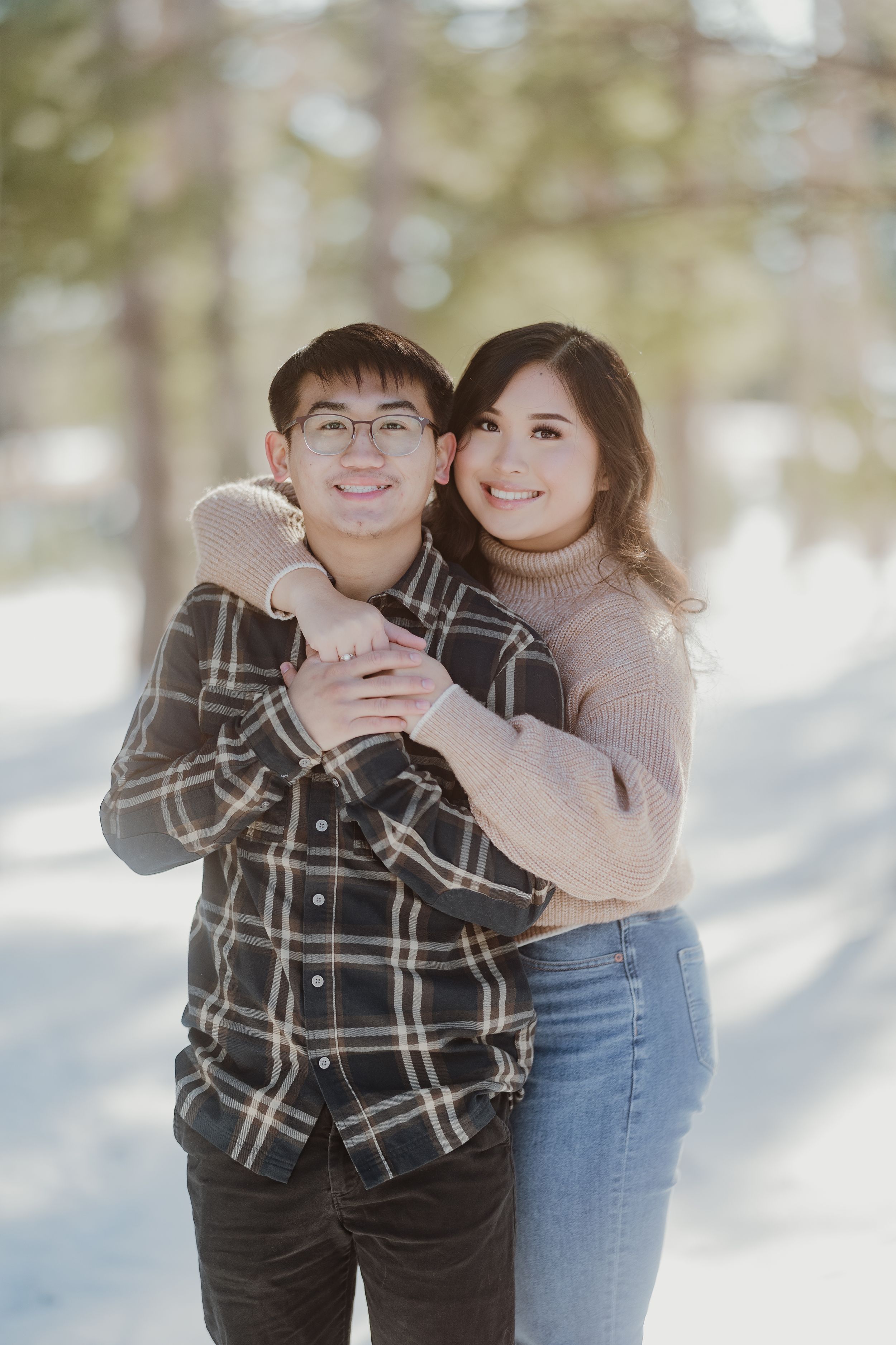Couple embraces while smiling in a bright, snow-covered winter forest setting.