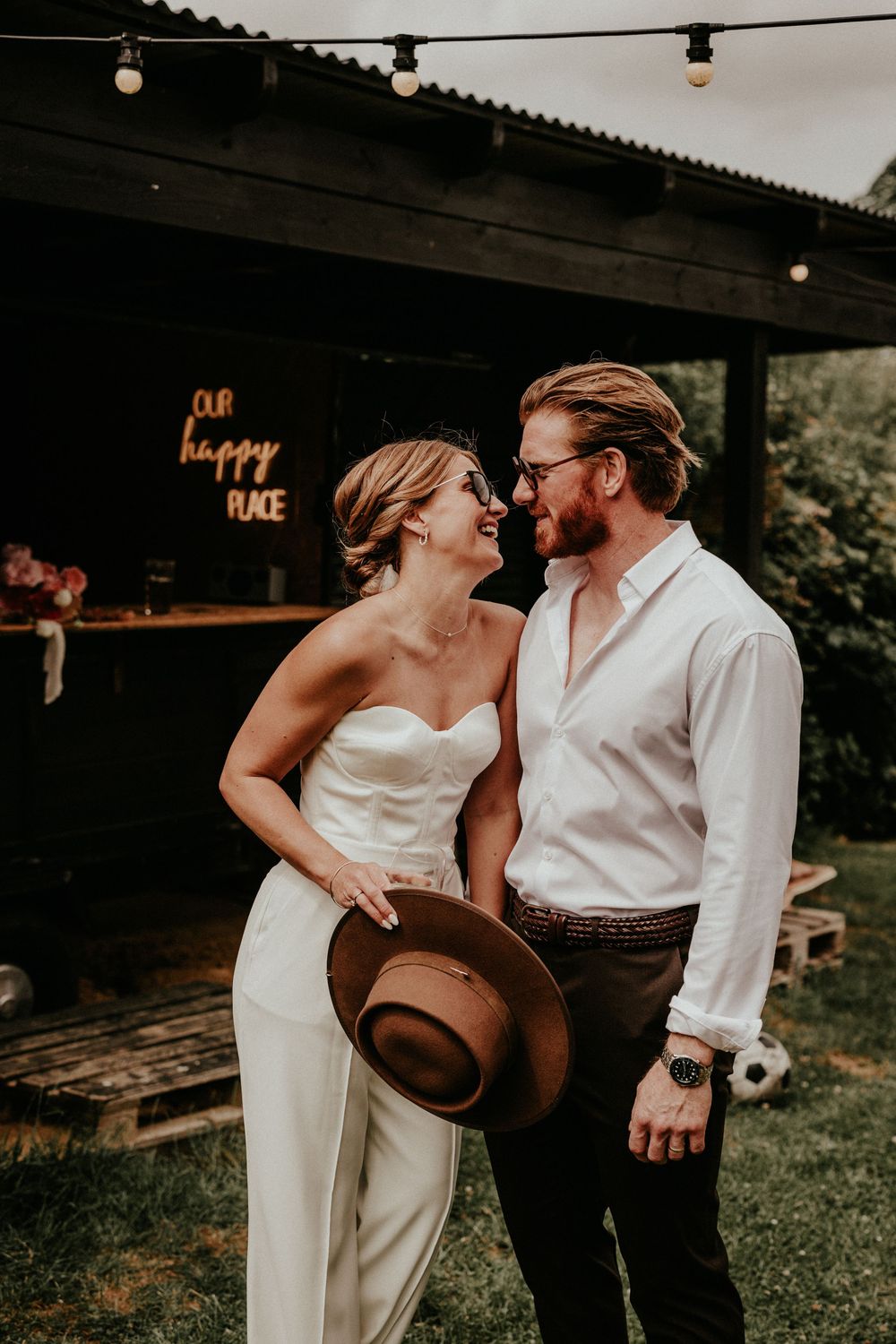 A romantic outdoor wedding moment at a rustic barn venue with string lights and a neon happy time sign glowing above.