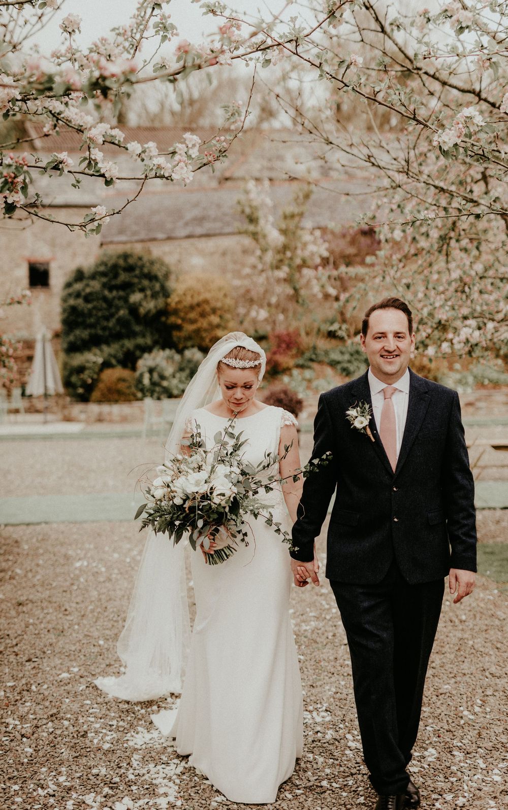 A bride and groom walk hand in hand under blooming cherry blossom trees on their wedding day.