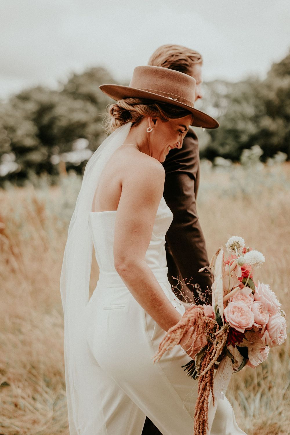 A romantic wedding couple walks together in a golden field while the bride carries a bouquet of pink roses and pampas grass.