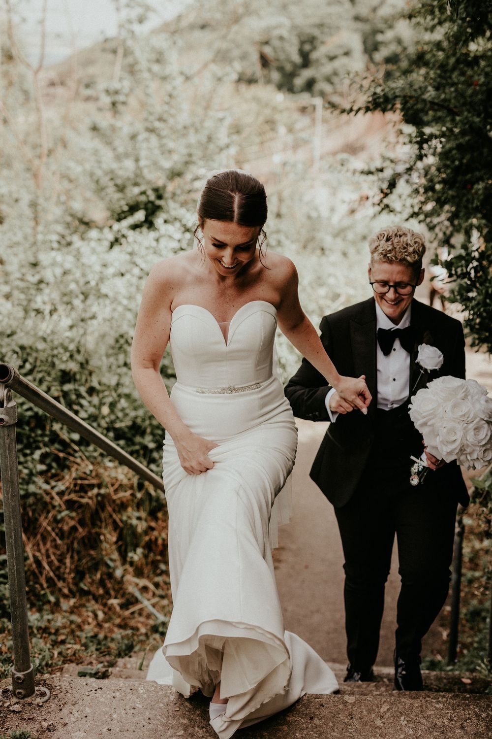 Two people in formal wedding attire carefully walk down outdoor stone steps surrounded by lush greenery.