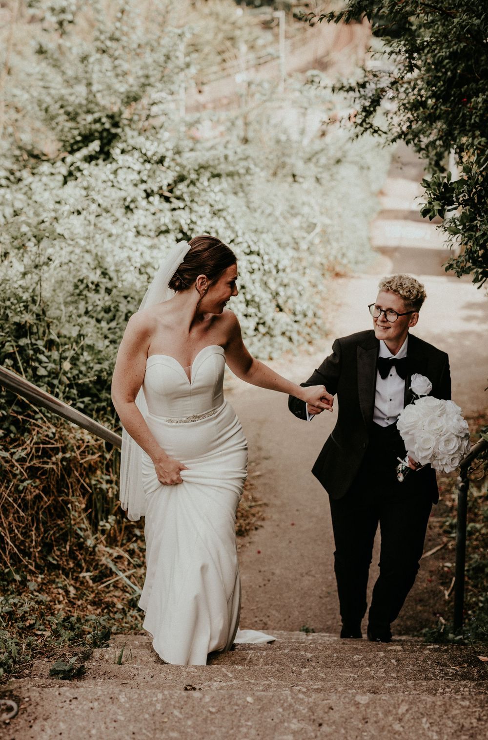 Bride in white gown and companion in black suit walking up rustic outdoor steps together at a wedding celebration.