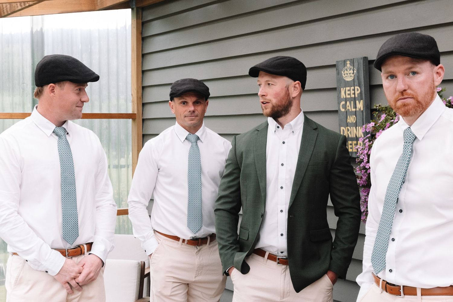 Groomsmen in matching flat caps, white shirts and khaki pants pose together in coordinated wedding attire.