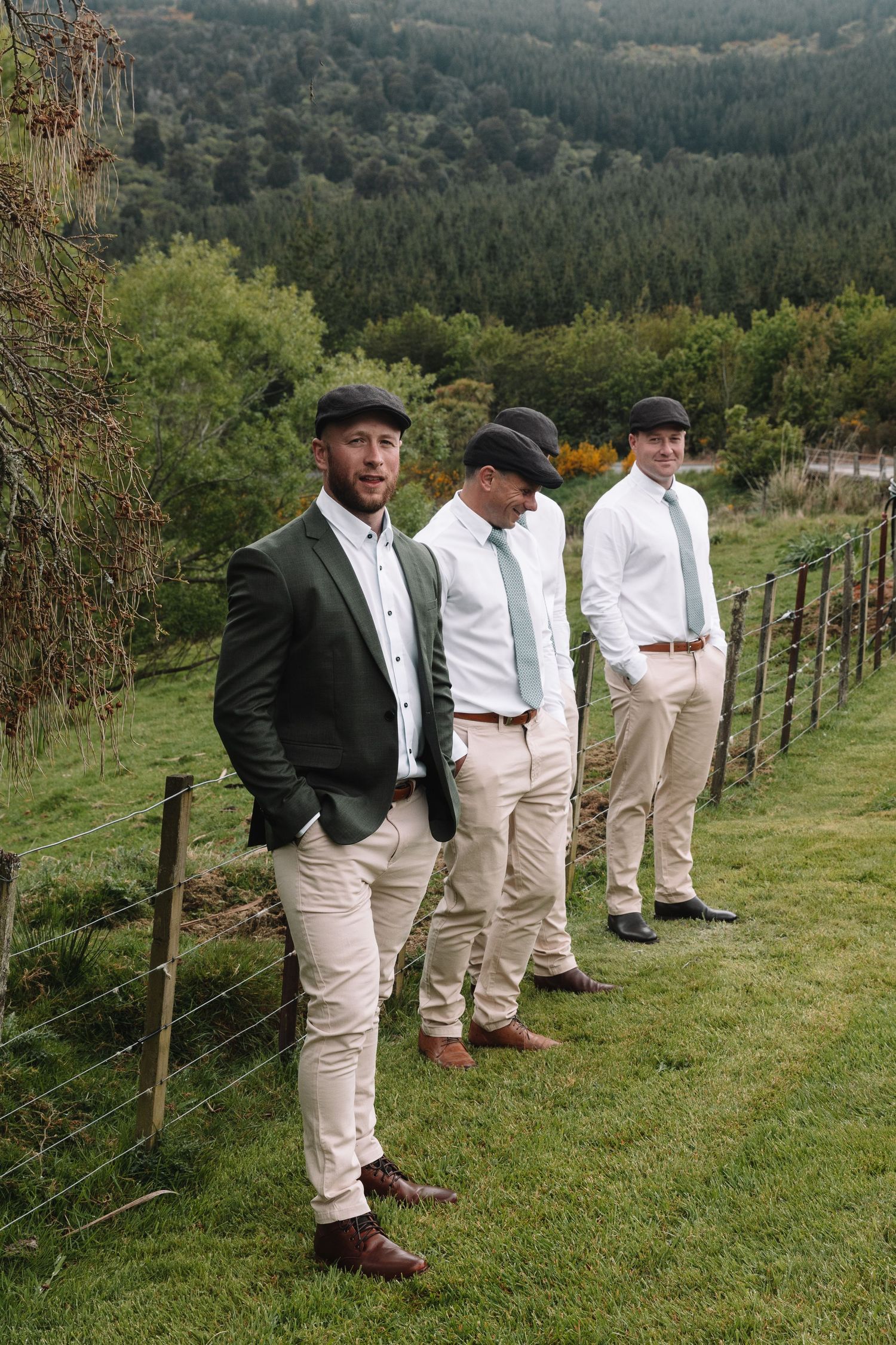 Wedding party members in flat caps and olive blazers stand on a hillside overlooking a forest landscape.