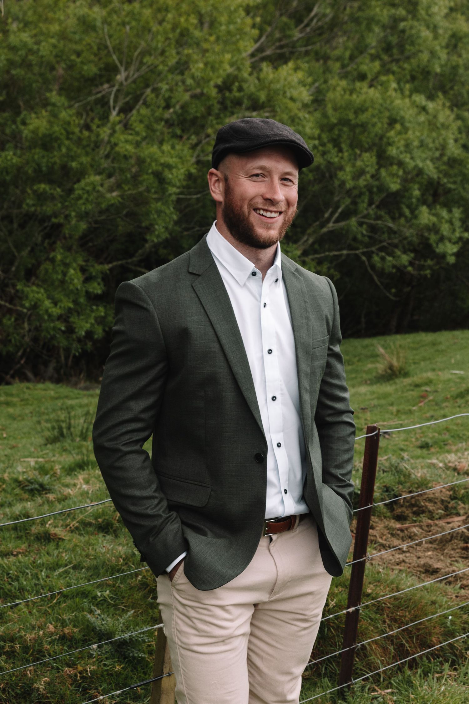 Wedding party member in olive blazer and flat cap poses against scenic mountain backdrop.