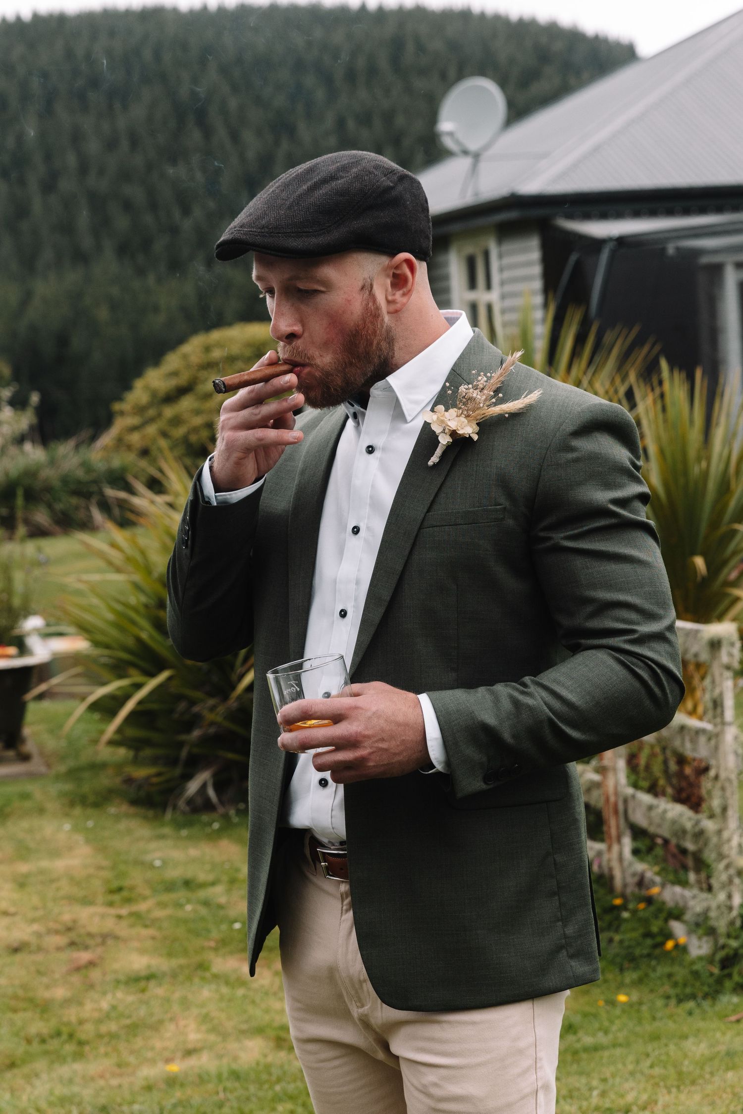 Wedding guest in olive blazer and flat cap enjoys a cigar and drink outdoors.