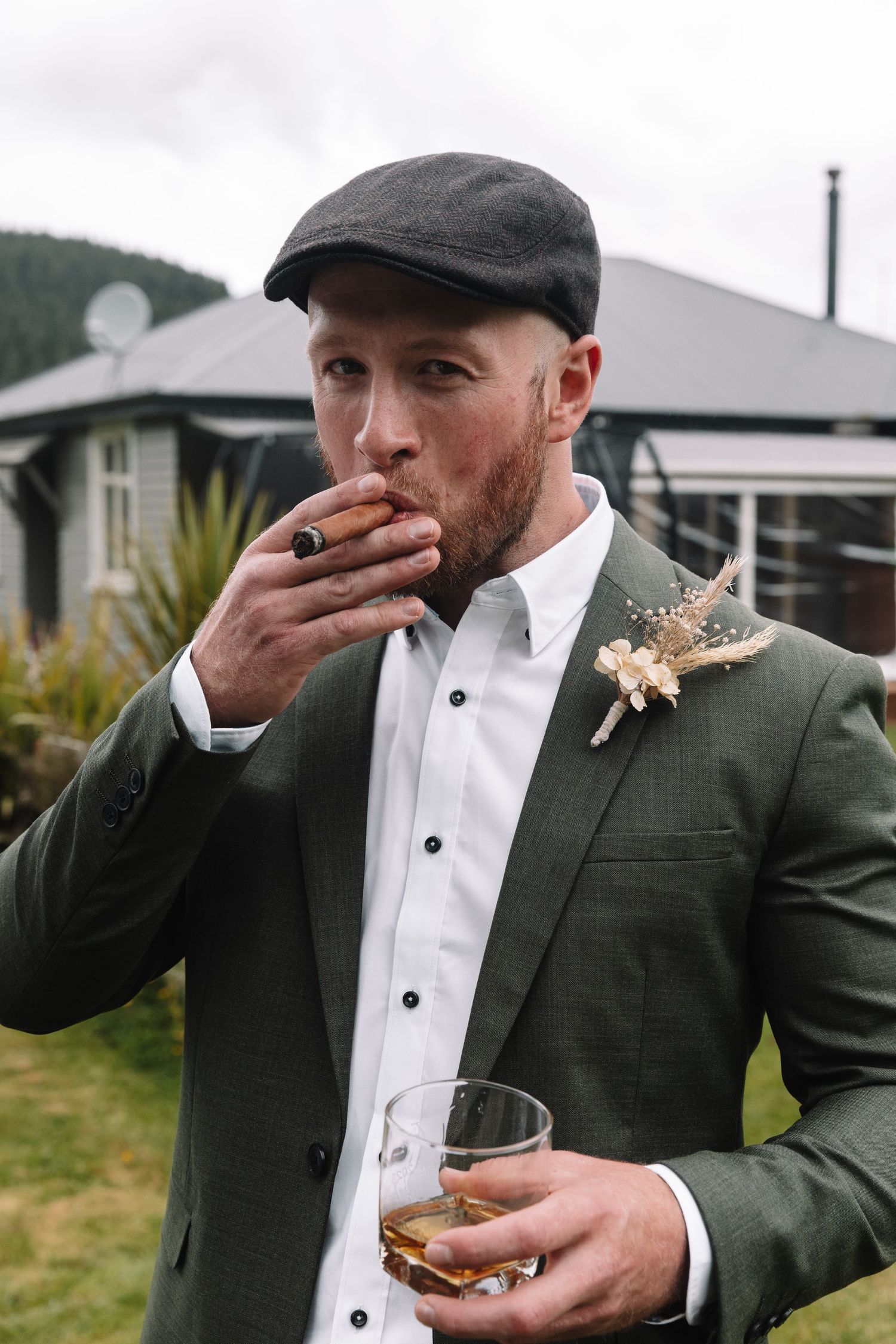 Wedding guest in olive blazer and flat cap enjoys cigar and drink against mountain backdrop.