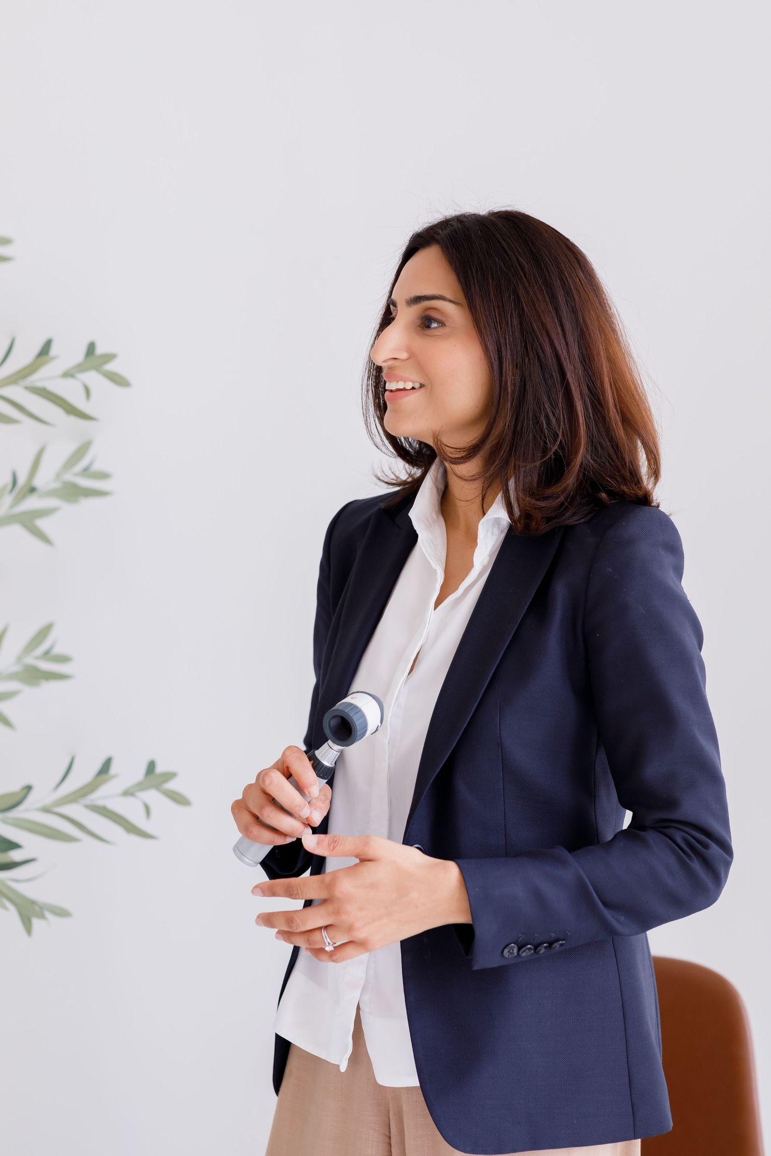 Professional speaker in navy blazer and white shirt holds microphone while presenting against light background with foliage decor.