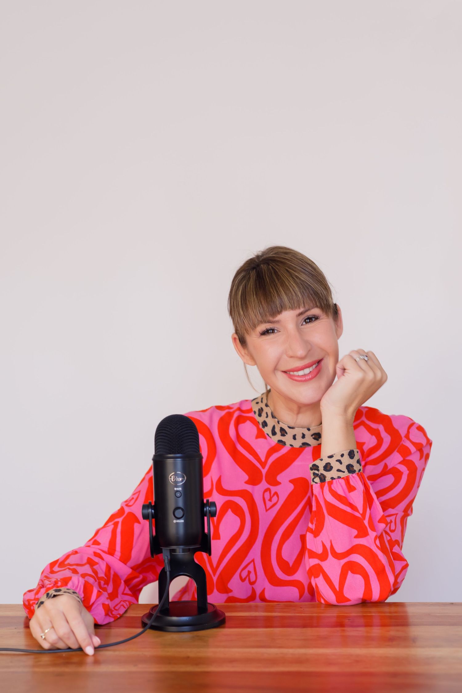 Professional podcaster sits at wooden desk with black microphone wearing bright pink and red patterned top and jewelry.
