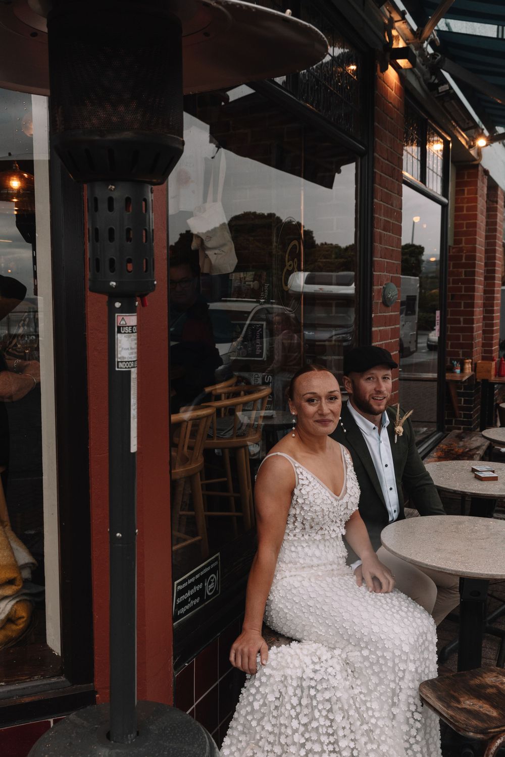 A couple sits at an outdoor cafe table dressed in formal wedding attire.