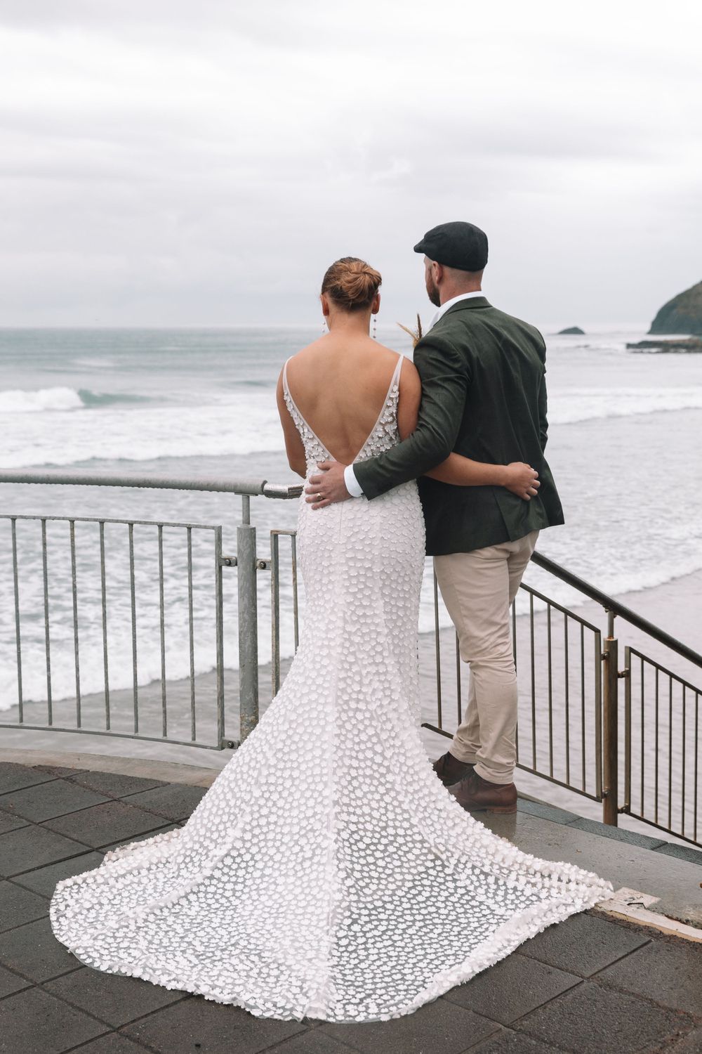 Newlywed couple enjoying ocean views from a deck while wearing wedding attire with bride in detailed lace gown.